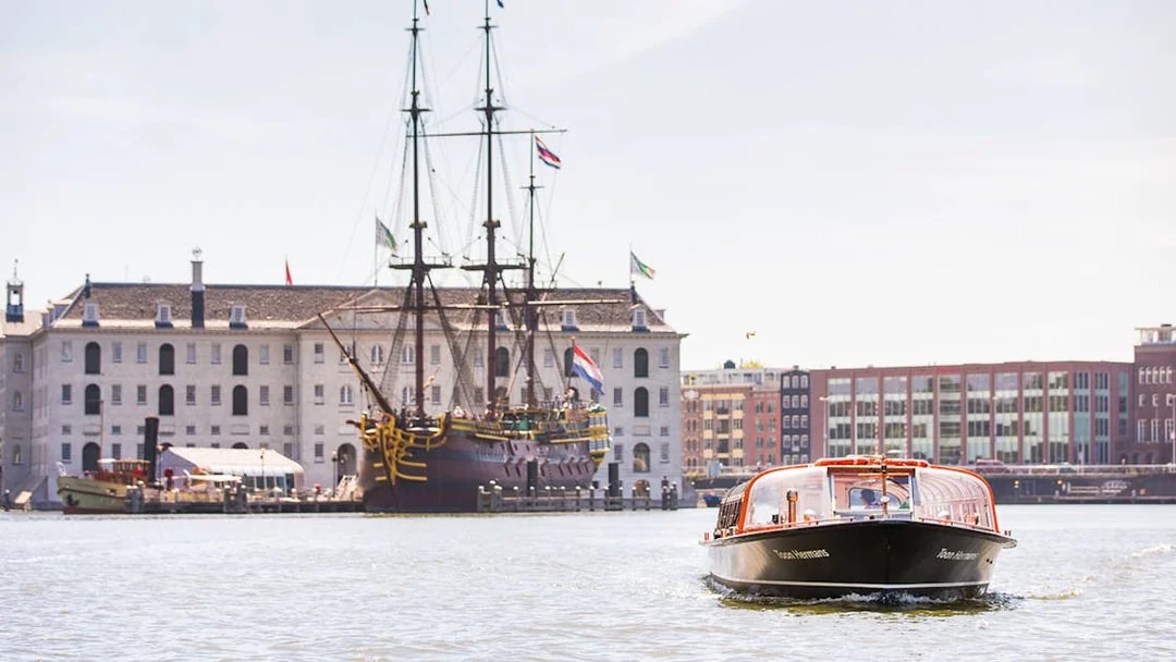 Amsterdam canal boat near the National Maritime Museum with historic ship in view.