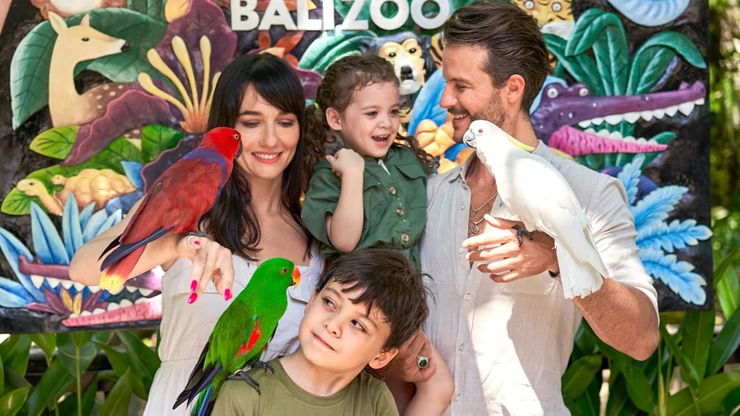 Family with colorful parrots in front of a mural at Bali Zoo.