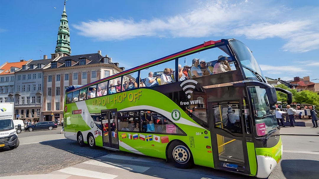 Hop-on hop-off bus in Copenhagen with tourists, near historic buildings and tower.