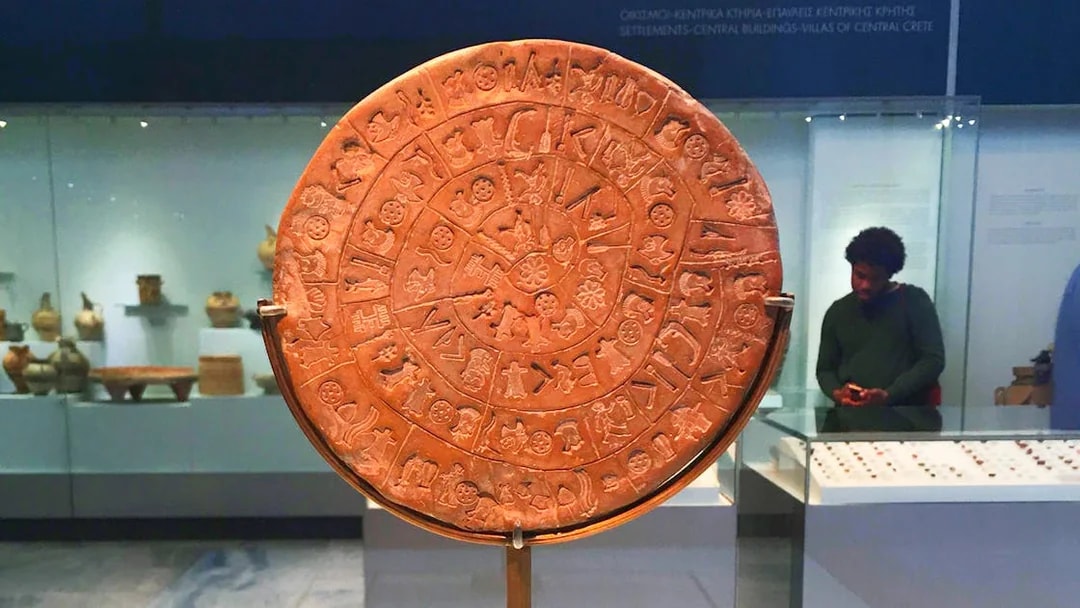 Phaistos Disc at Heraklion Archaeological Museum, Crete, with visitors in the background.