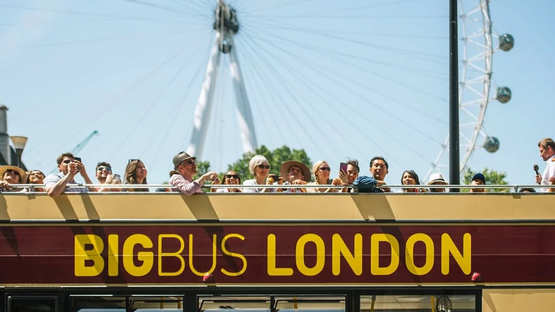 Open-top Big Bus tour in London with tourists, London Eye in background.