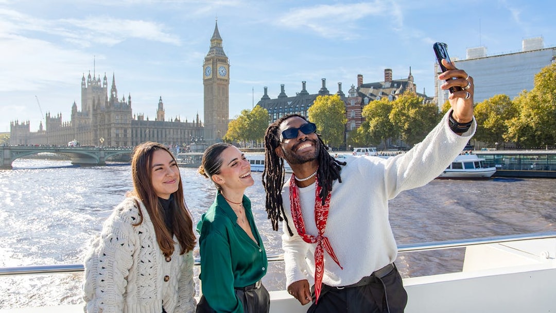 Friends taking a selfie on a Thames River cruise with Big Ben in the background, London.