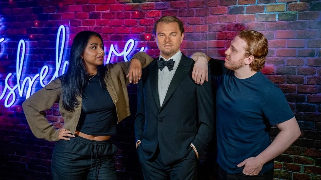 Wax figure of a man in a tuxedo at Madame Tussauds, London, with two visitors posing beside it.