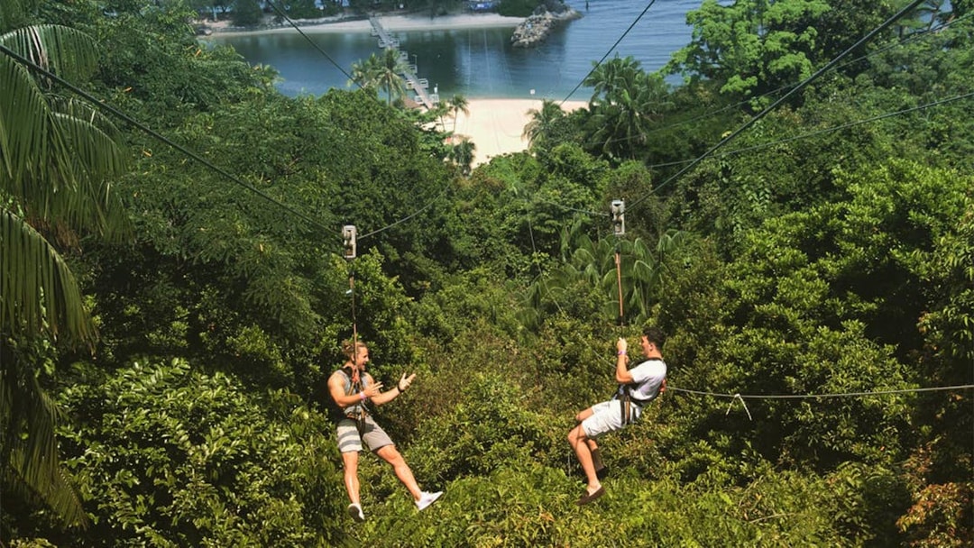 Two people ziplining over lush greenery towards a beach in Singapore.