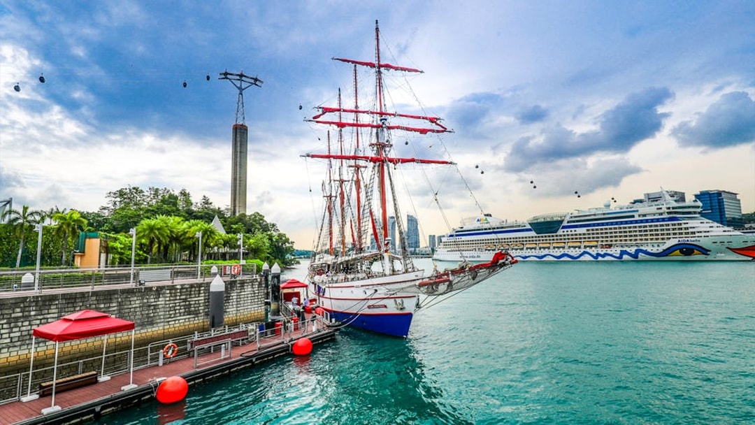 Tall ship docked at Singapore harbor with cable cars and cruise ship in the background.