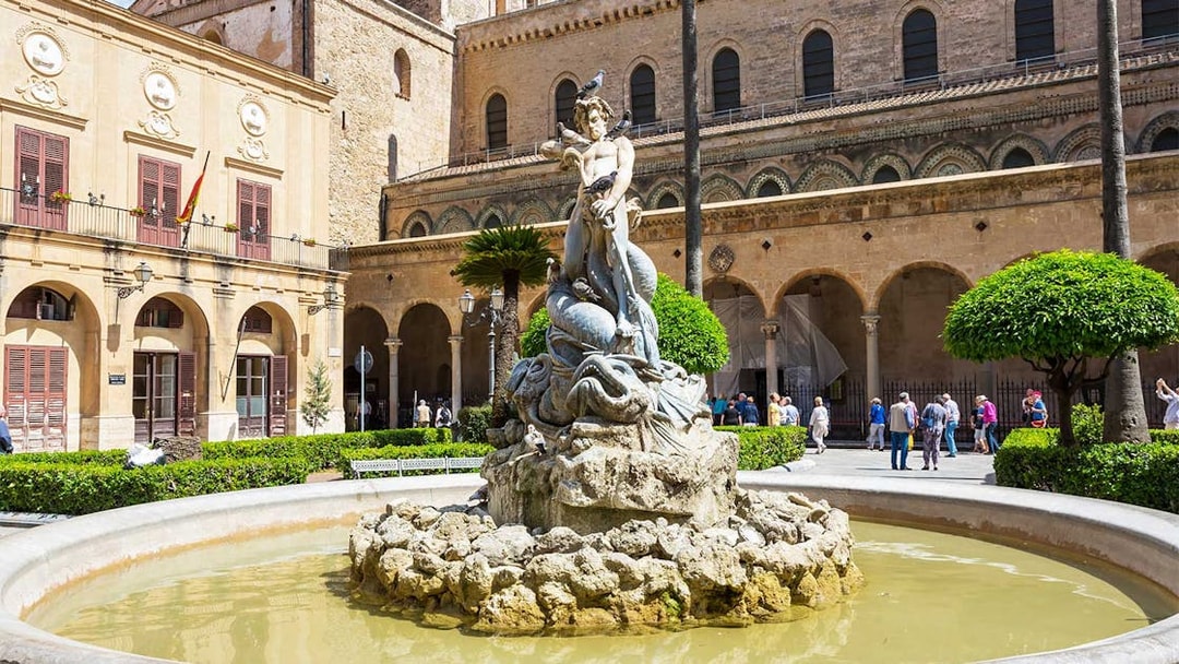 Fountain with statue in Monreale square, Sicily, with historic architecture in the background.