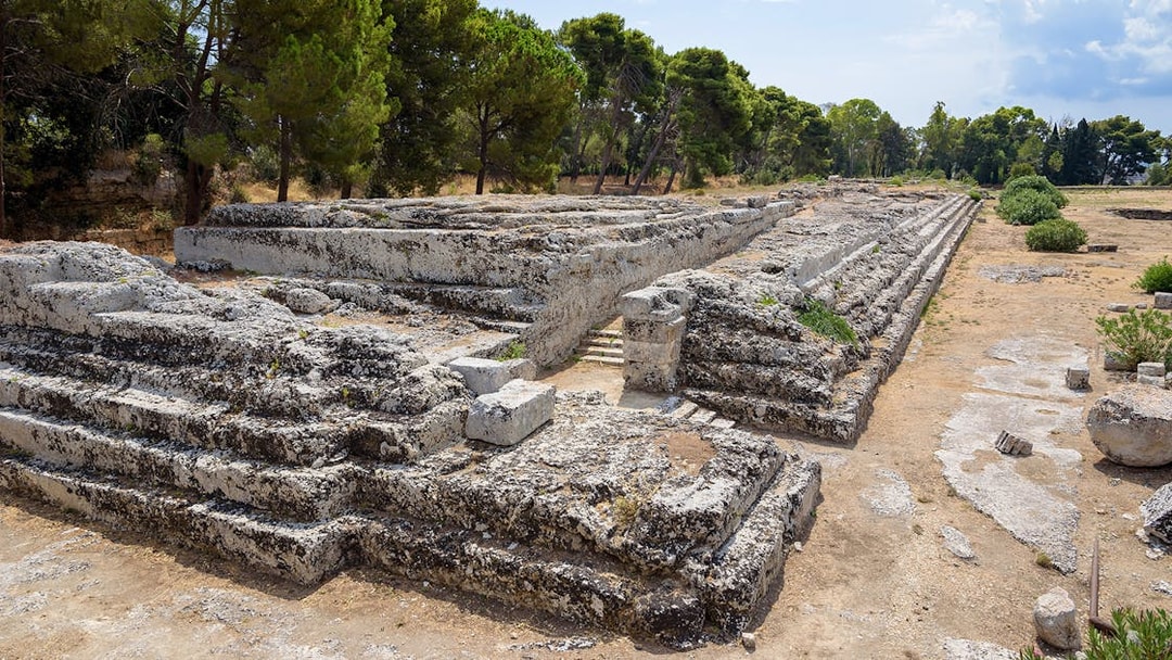 Ancient stone ruins at Neapolis Archaeological Park, Syracuse, surrounded by trees.