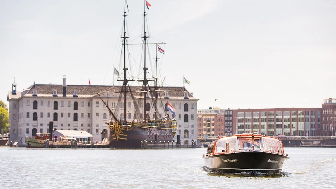 Amsterdam canal cruise boat near NEMO Science Museum with historic ship in background.