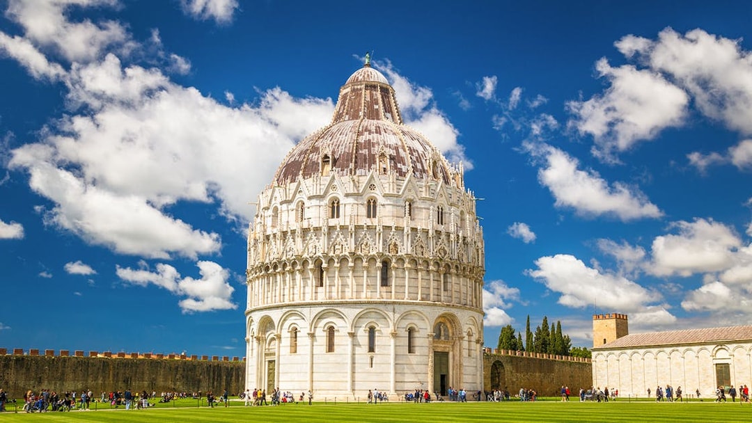 Pisa Baptistery with tourists on green lawn under blue sky.