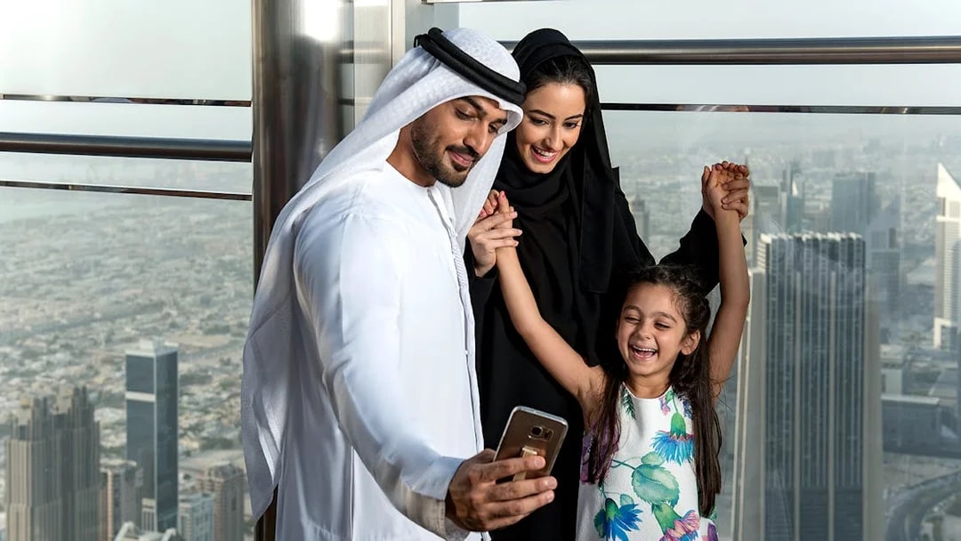 Family taking a selfie at Burj Khalifa observation deck, Dubai skyline in background.