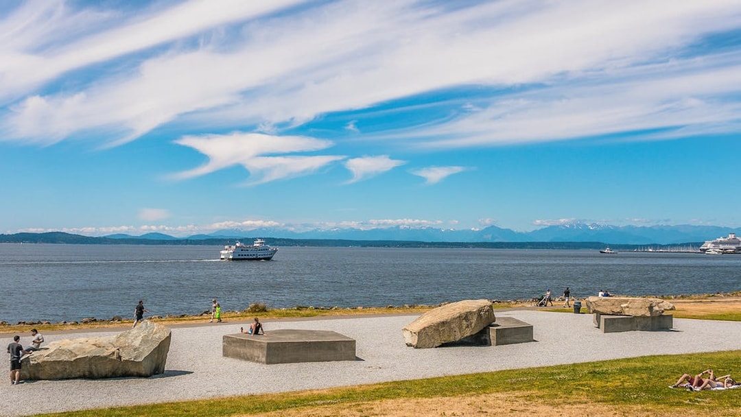 Seattle waterfront view with ferry and people relaxing near sculptures.