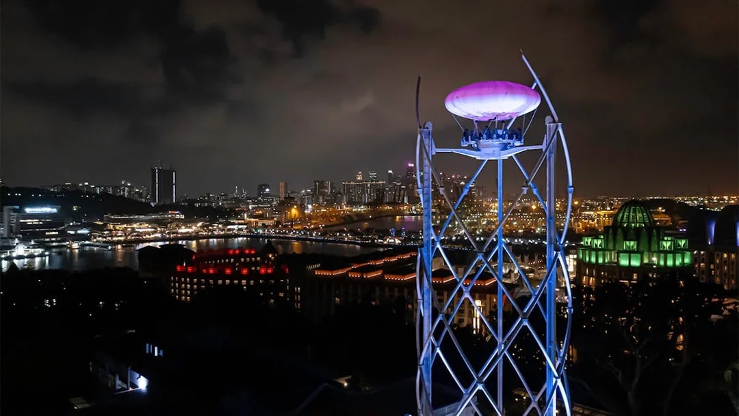 SkyHelix Sentosa ride illuminated at night with Singapore cityscape in the background.