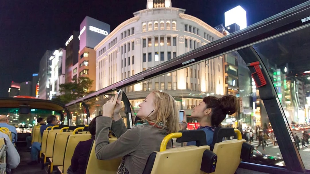 Open-top bus tour in Tokyo at night, passengers viewing illuminated Ginza buildings.