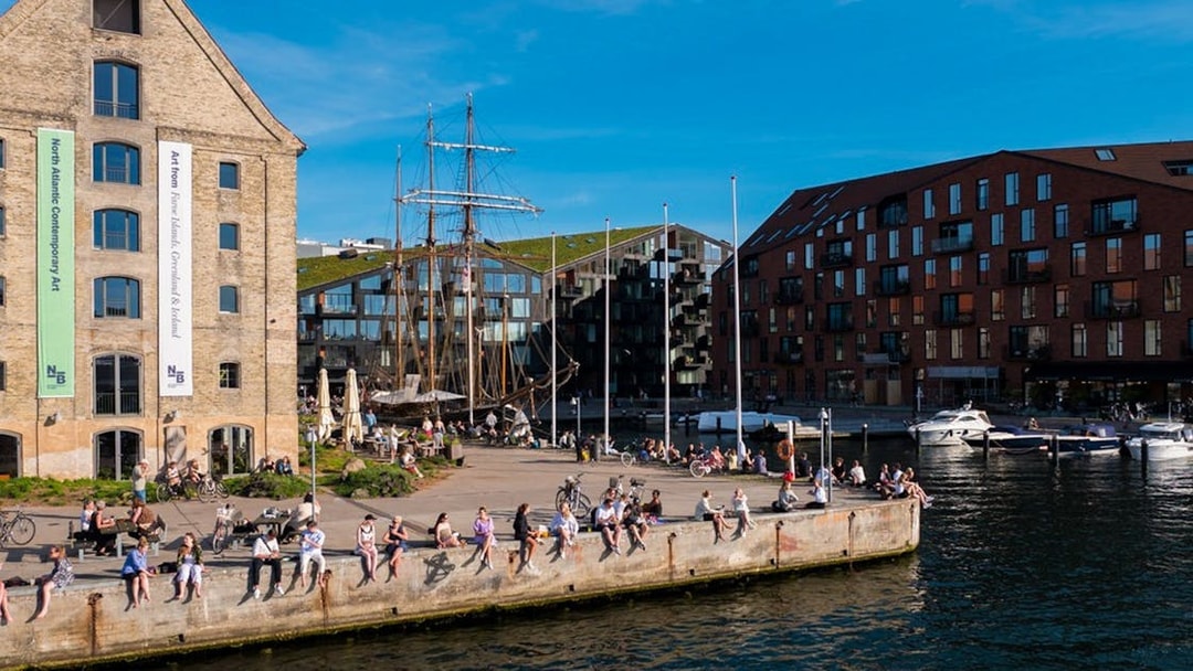 People relaxing by the waterfront in Copenhagen with historic buildings and boats in view.