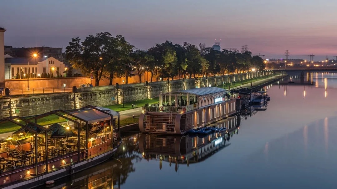 Krakow riverside at dusk with illuminated boats and tree-lined promenade.