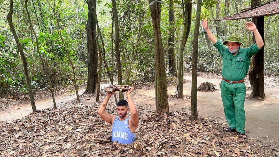 Man emerging from Cu Chi Tunnel holding cover, with guide nearby in forest setting.