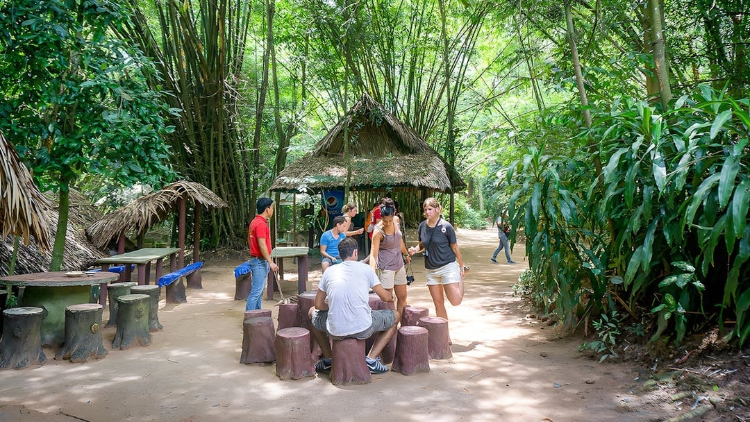 Visitors gathering at Cu Chi Tunnels, Vietnam, near a thatched hut surrounded by lush greenery.