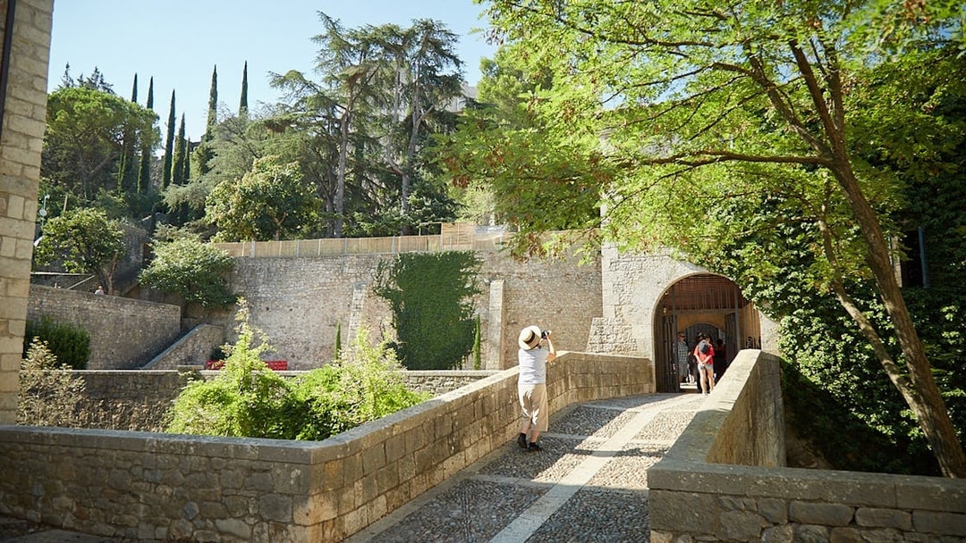Stone bridge leading to historic entrance in Girona, surrounded by lush greenery.