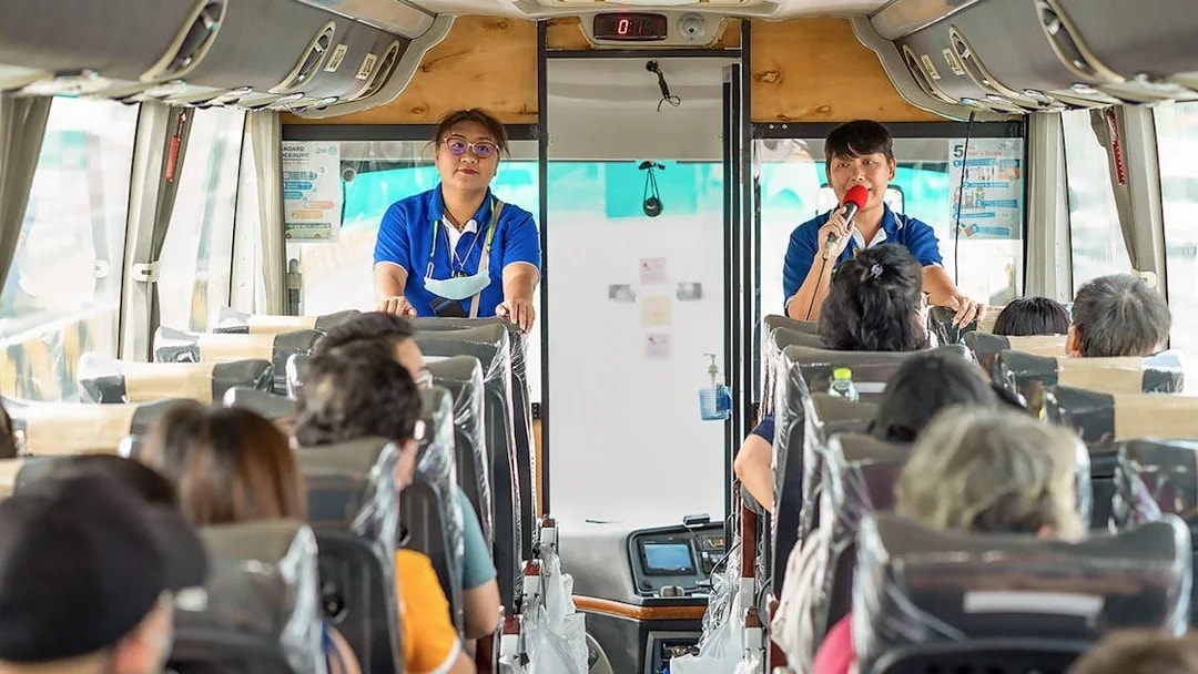 Tour guide speaking to passengers on a bus for Damnoen Saduak Floating Market tour.