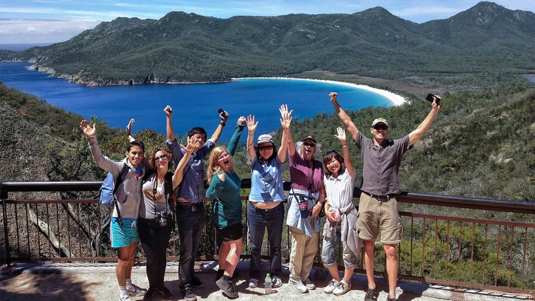 Tour group celebrating at Wineglass Bay lookout with ocean and mountains in background.