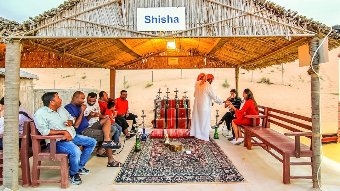 Shisha lounge in desert safari with people enjoying hookah under a thatched roof.