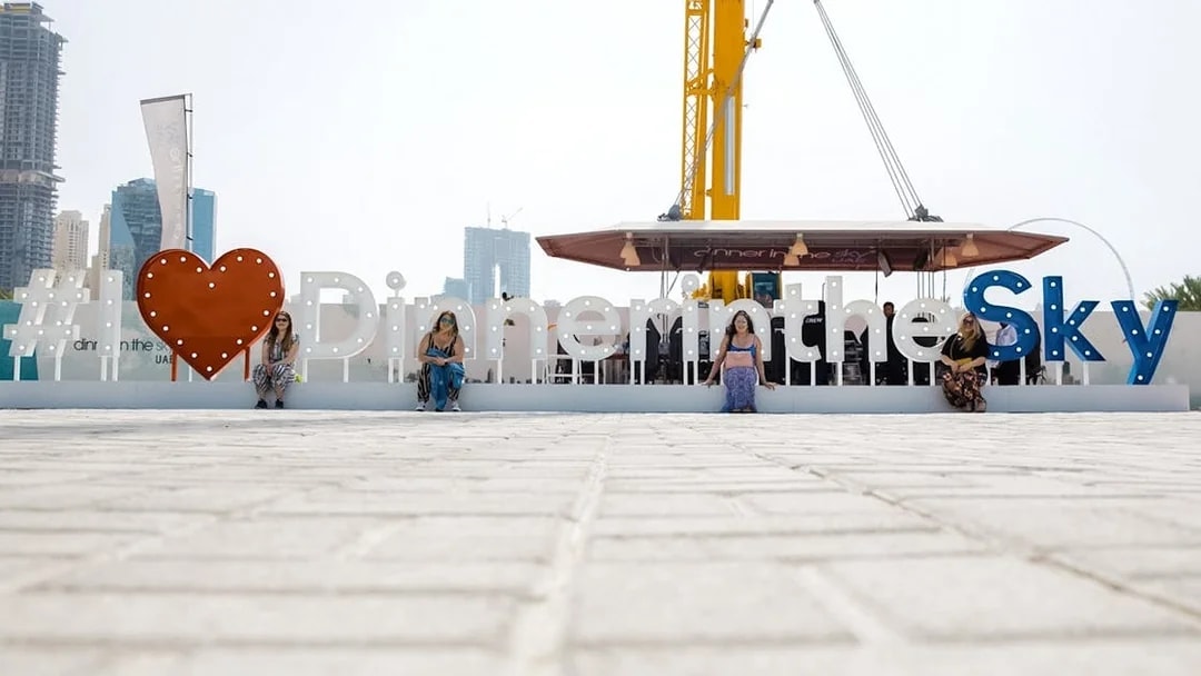 Dinner in the Sky sign with people seated, crane visible, city skyline in background.