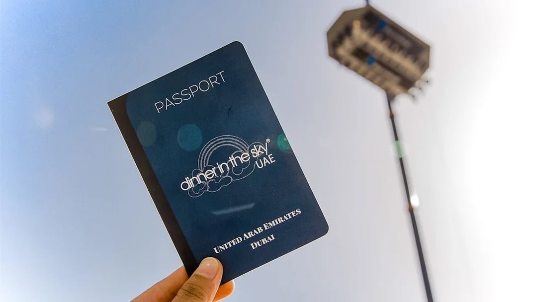 Hand holding a Dinner in the Sky UAE passport with a suspended dining platform in the background.