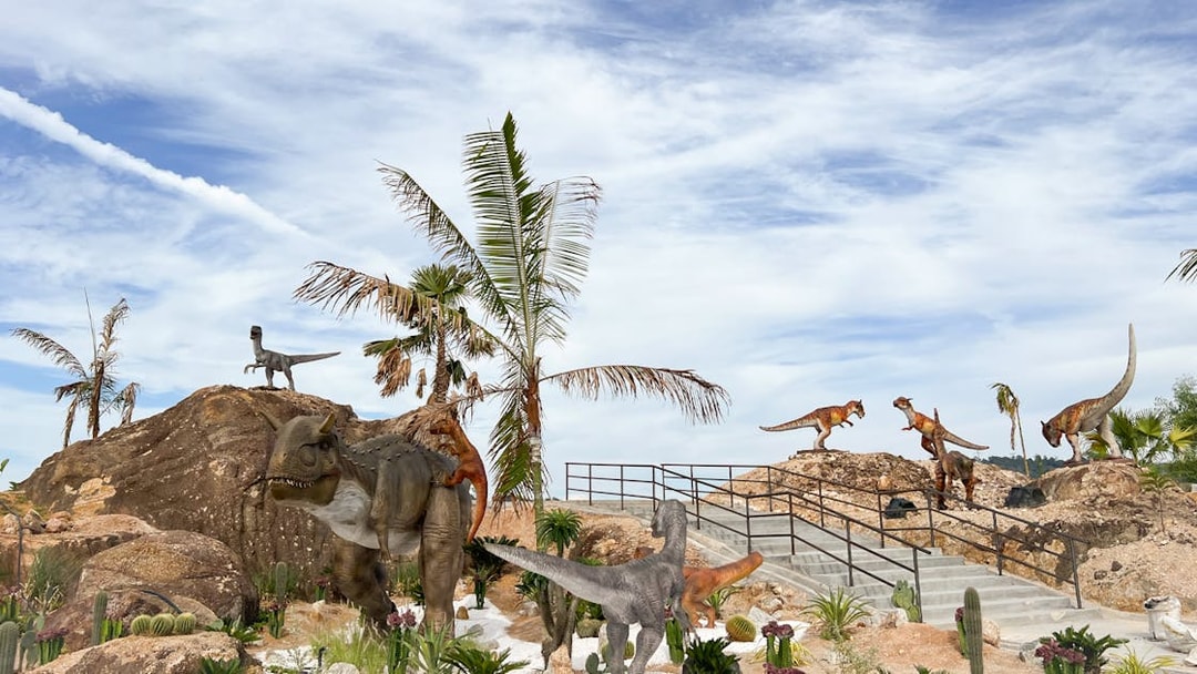 Dinosaurs in a desert landscape with palm trees and rocky terrain, Dino Desert Ticket.