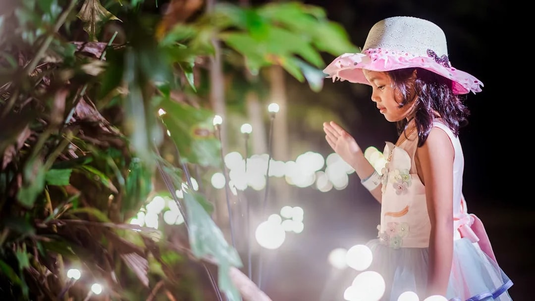 Little girl in a hat admiring glowing lights in a garden setting.