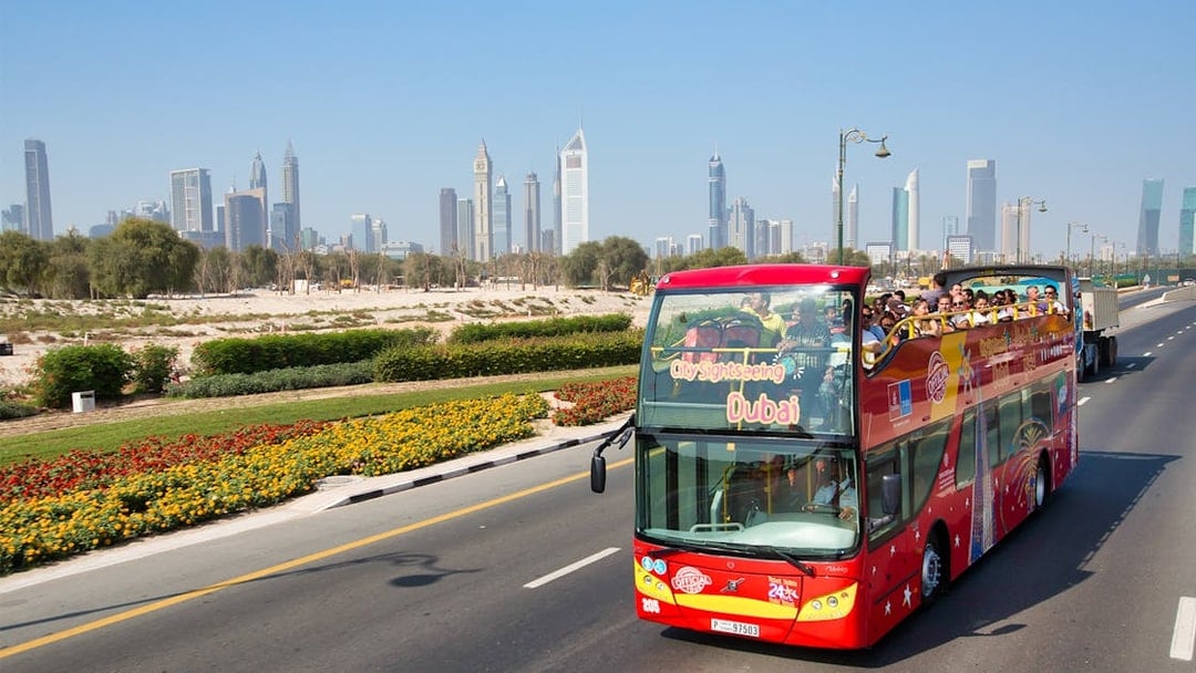 Open-top tour bus on Dubai road with city skyline in background.