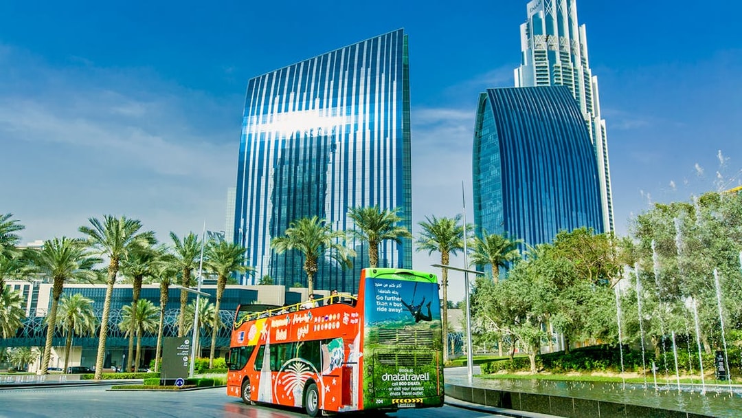 Hop-on-hop-off bus in front of modern skyscrapers and palm trees in Dubai.