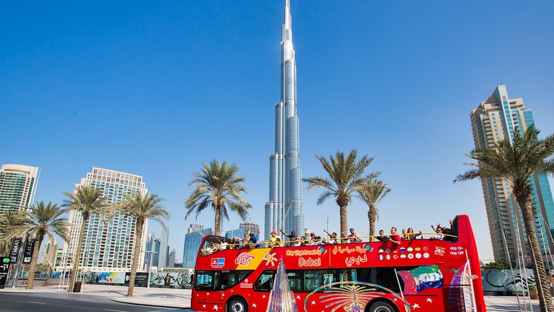 Open-top tour bus near Burj Khalifa, Dubai, with tourists enjoying the view.