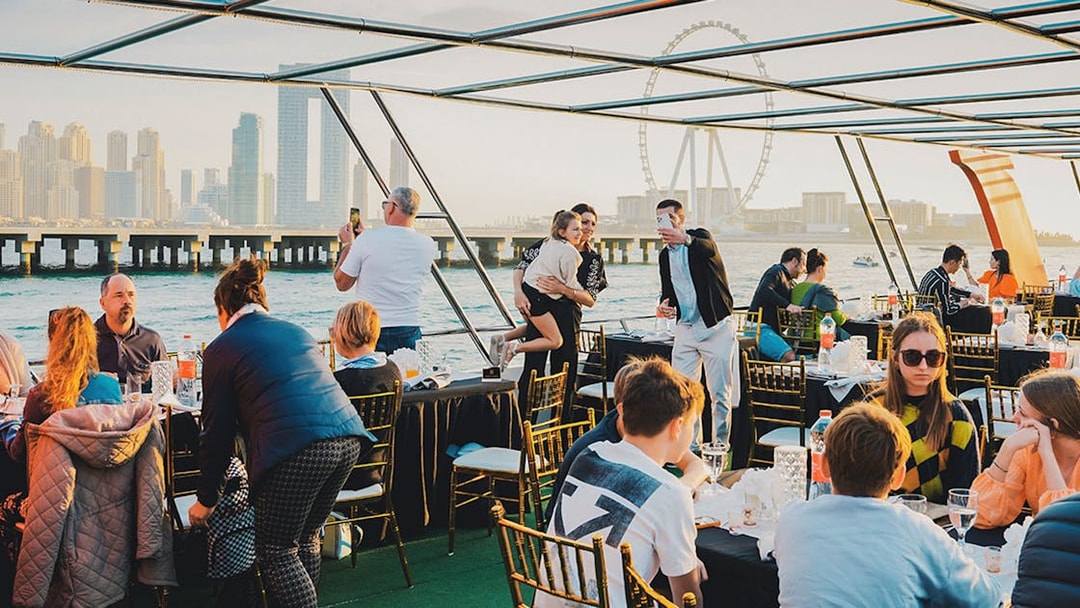 Guests dining on a sunset cruise in Dubai Marina with city skyline and Ferris wheel in view.