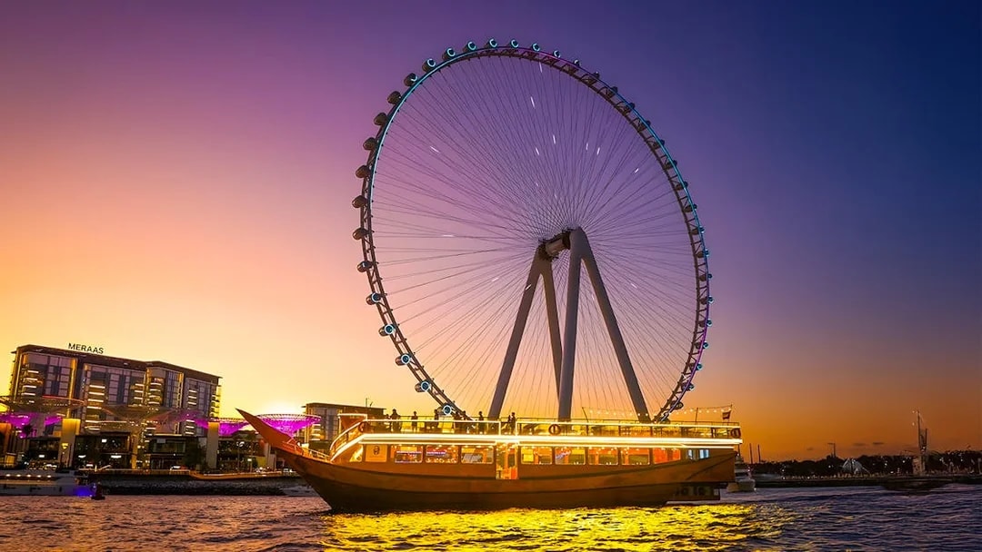 Dhow cruise with lights on Dubai Marina at sunset, near Ferris wheel.