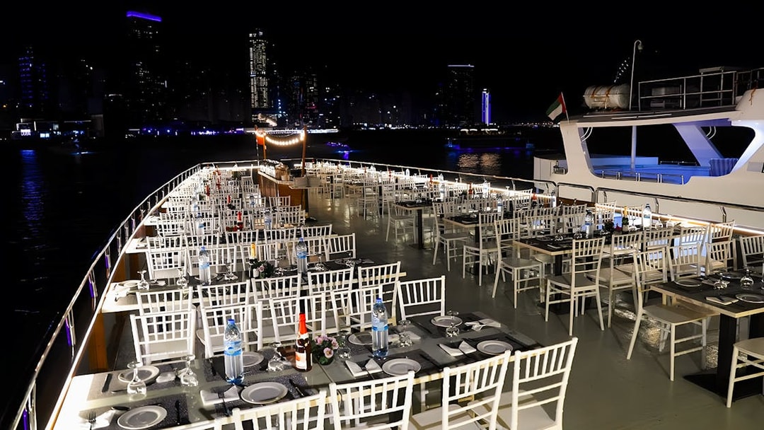 Dhow cruise dinner setup with tables and chairs on deck, Dubai Marina skyline at night.