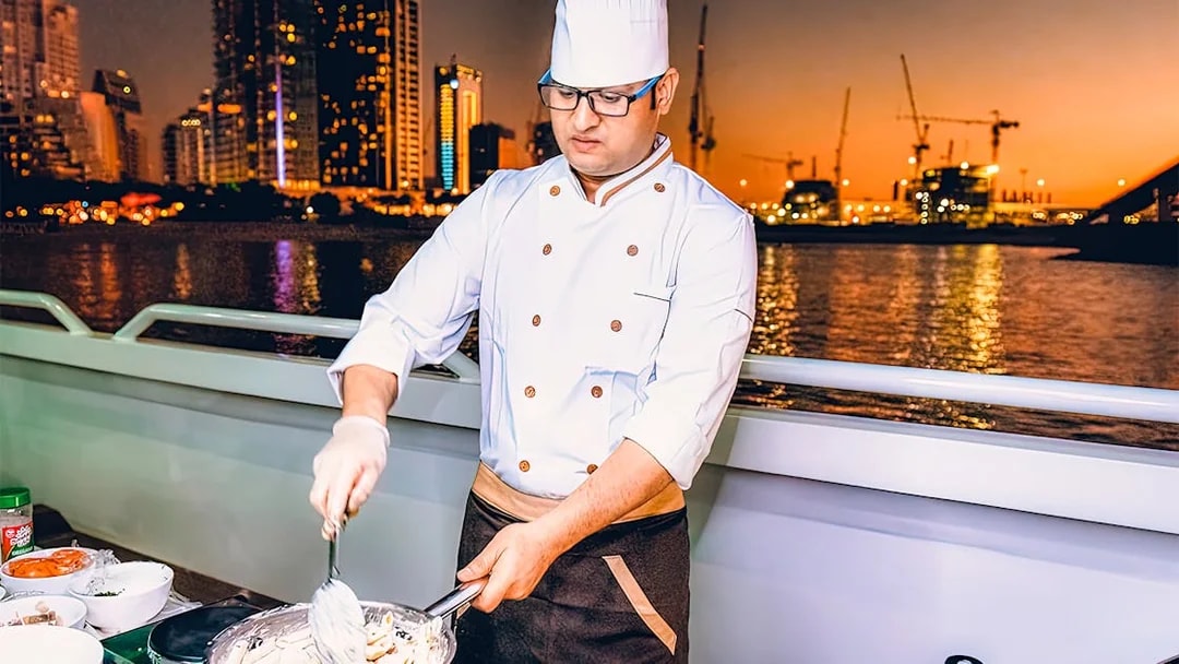 Chef preparing meal on Mega Yacht Dinner Cruise in Dubai with city skyline at sunset.