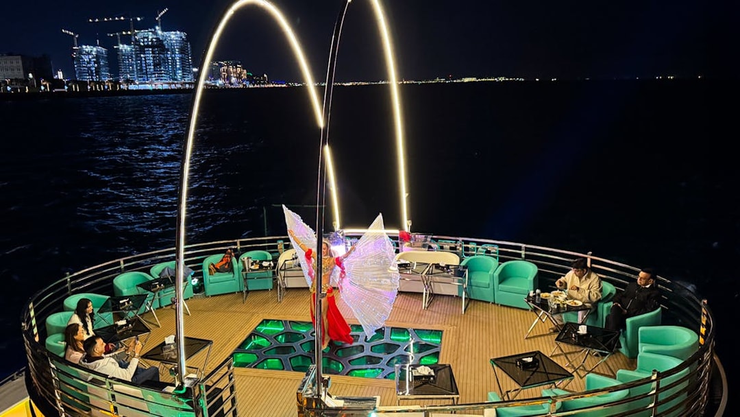 Performer with illuminated wings on a nighttime cruise deck, city lights in the background.