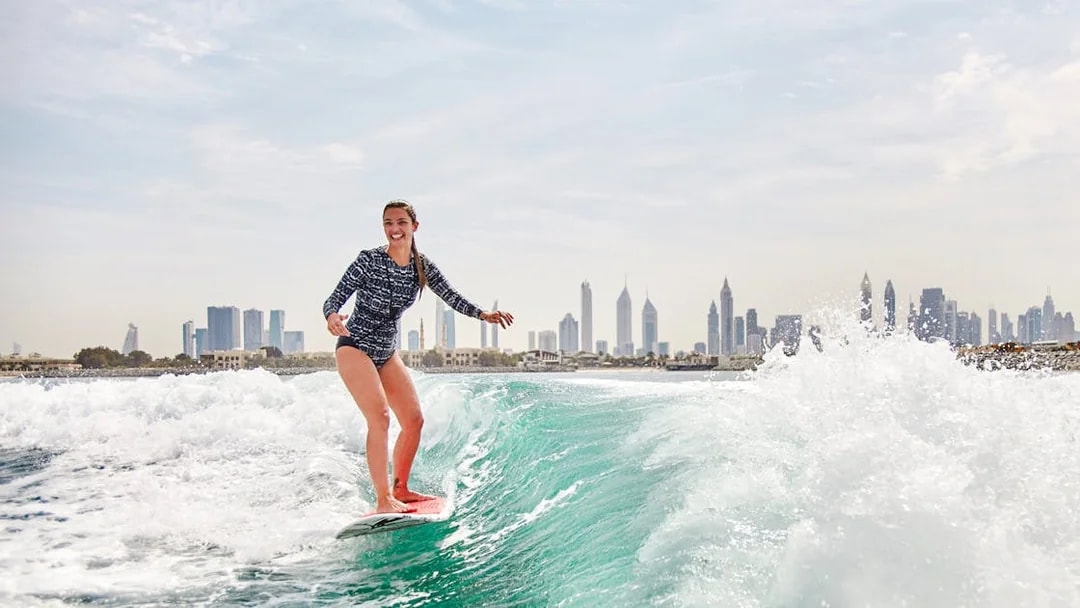 Surfer riding a wave with Dubai skyline in the background.