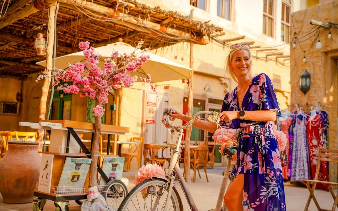 Woman with bicycle in front of a coffee shop with floral decor in a vibrant marketplace.