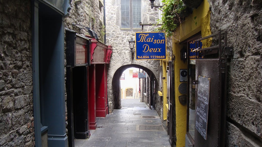 Narrow stone alleyway in Kilkenny with colorful shop signs and archway.