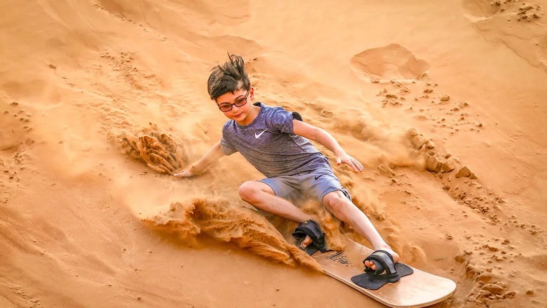 Young person sandboarding down a dune during a desert safari in Dubai.