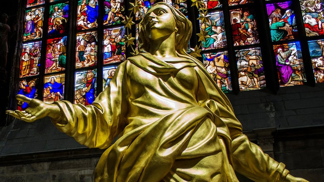 Golden statue inside Milan Cathedral with colorful stained glass windows in the background.