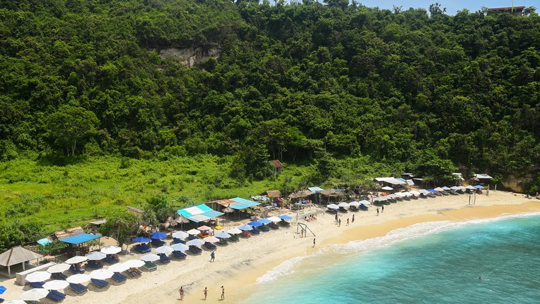 Beach with umbrellas and tourists at Nusa Penida Island, Indonesia, surrounded by lush greenery.