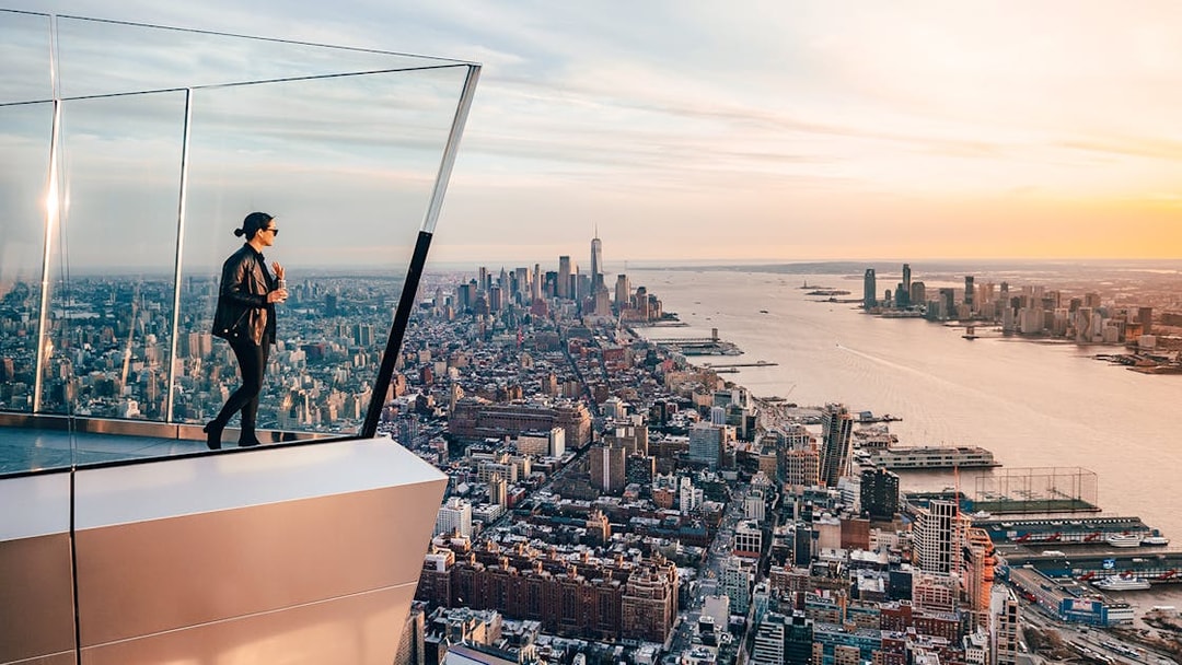 Person standing on The Edge observation deck overlooking New York City skyline at sunset.