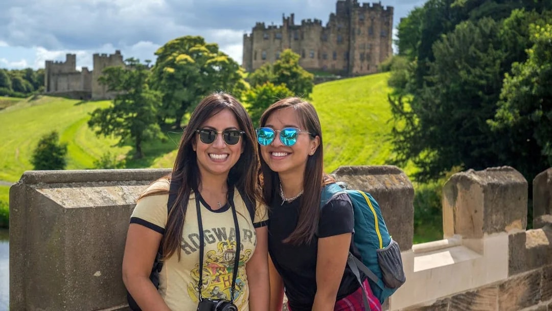 Two tourists smiling with Alnwick Castle in the background on a day tour from Edinburgh.