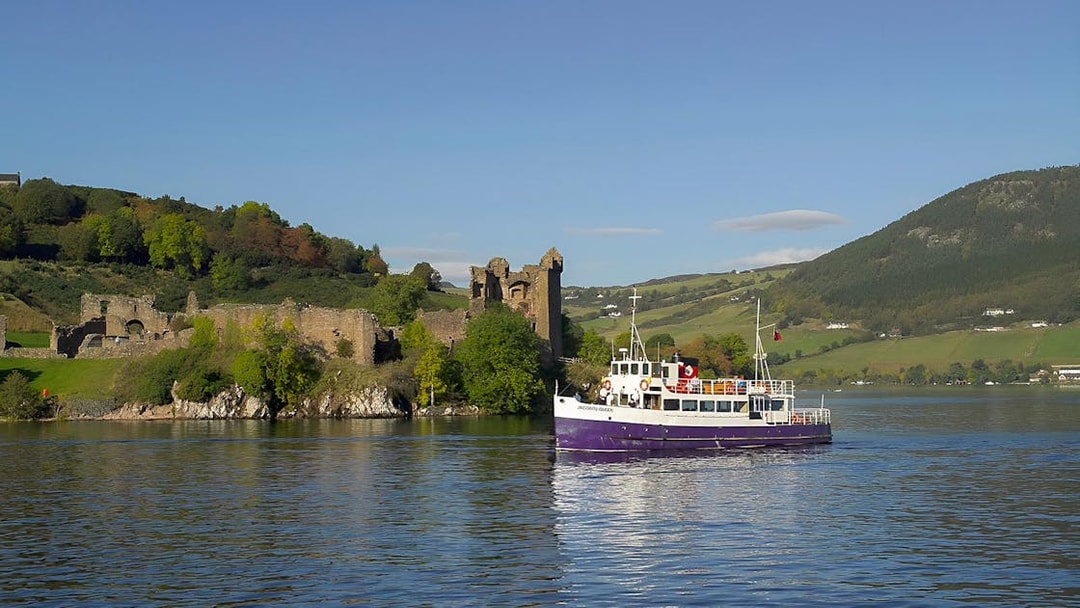 Boat cruising near Urquhart Castle on Loch Ness, Scotland, with green hills in the background.