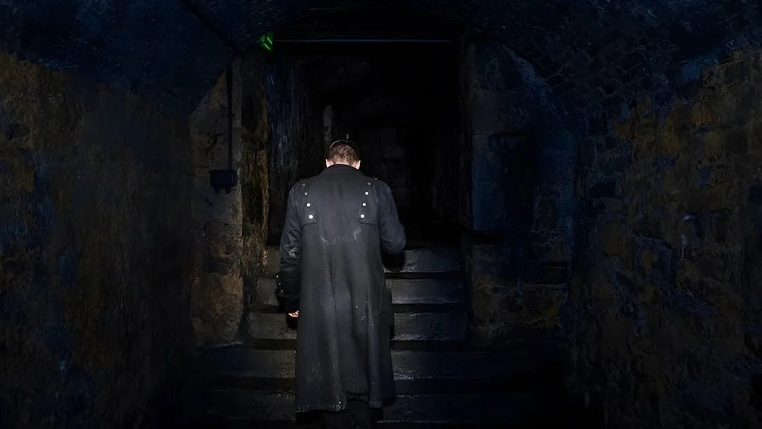 Man in dark coat ascending stone steps in dimly lit underground passage.