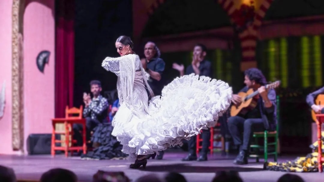 Flamenco dancer in white dress performing on stage with musicians in background.