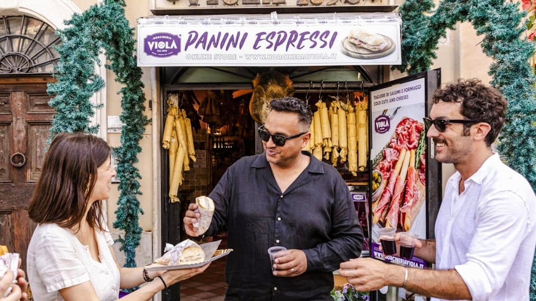 Guide serving panini to guests at a Rome food tour outside a deli.