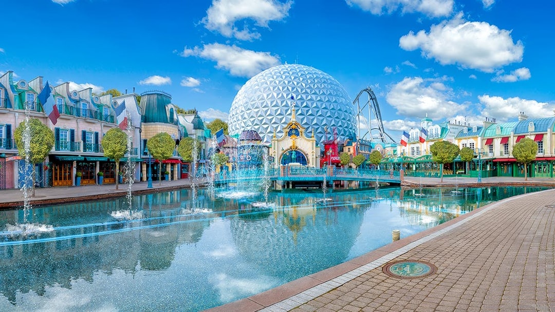 Theme park with geodesic dome, fountains, and colorful buildings reflecting in water.